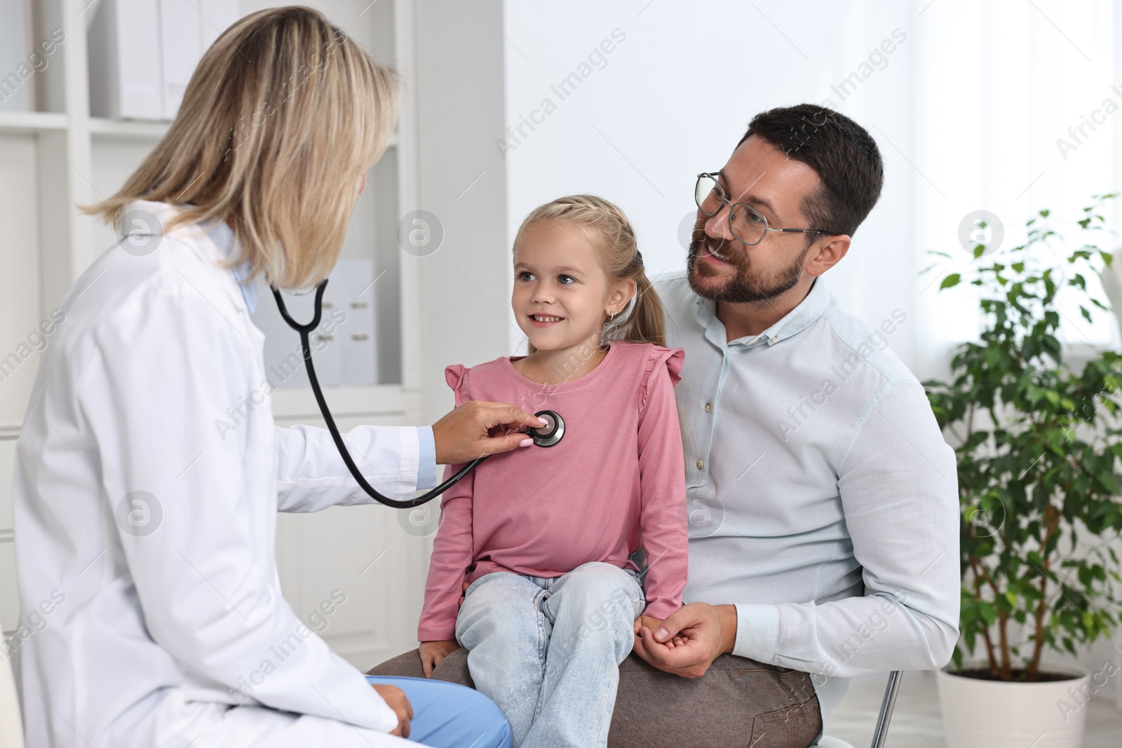 Photo of Doctor examining little girl with stethoscope and her father in hospital