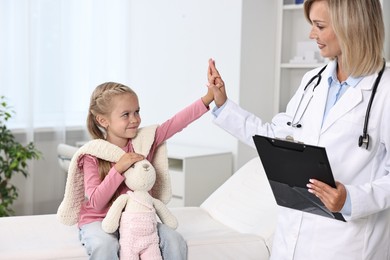 Photo of Doctor with clipboard giving high five to little girl with toy in hospital