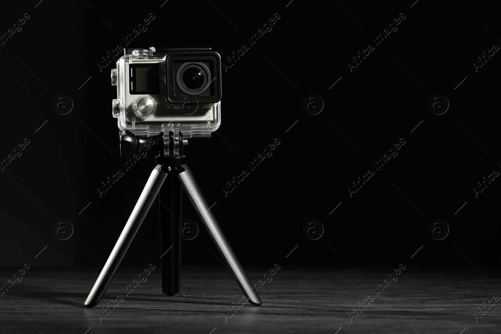 Photo of Modern action camera and tripod on wooden surface against black background. Space for text