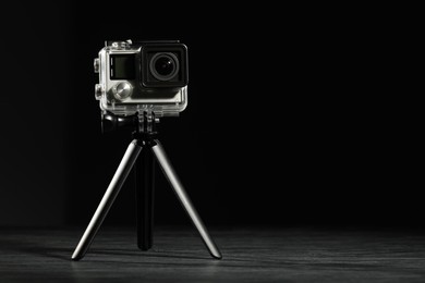 Modern action camera and tripod on wooden surface against black background. Space for text