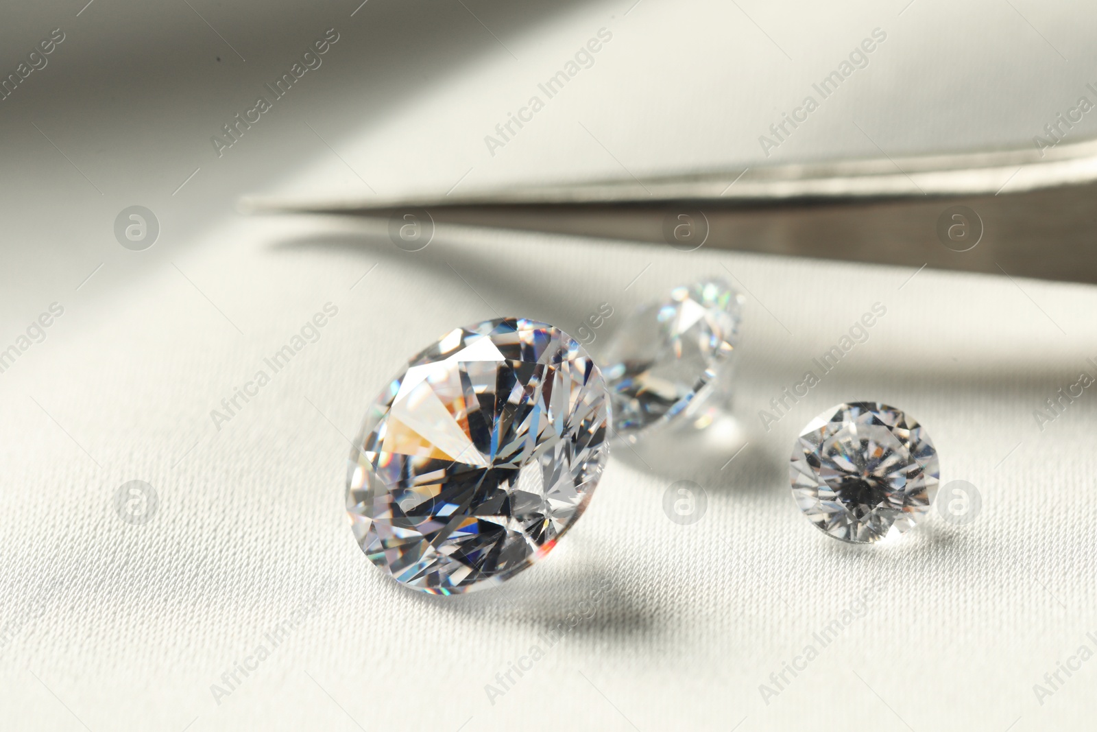 Photo of Beautiful shiny diamonds and tweezers on white fabric, closeup