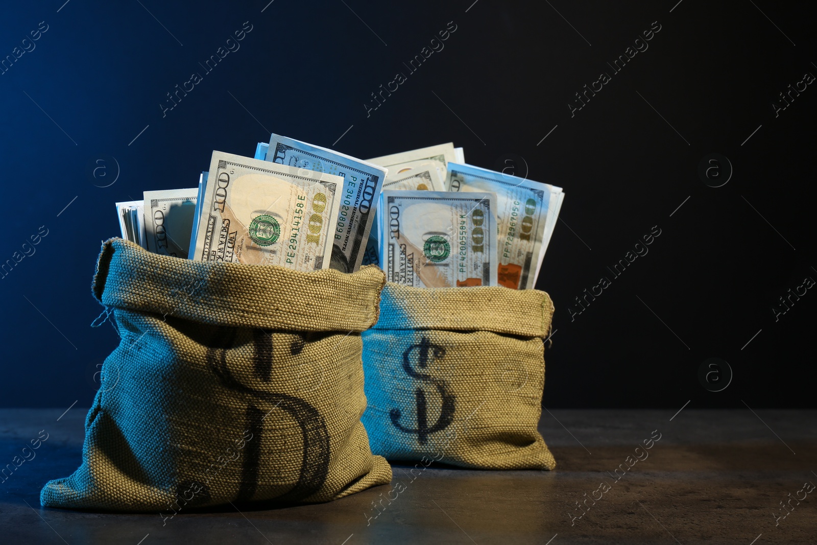 Photo of Dollar banknotes in burlap sacks on grey table against dark background in neon lights