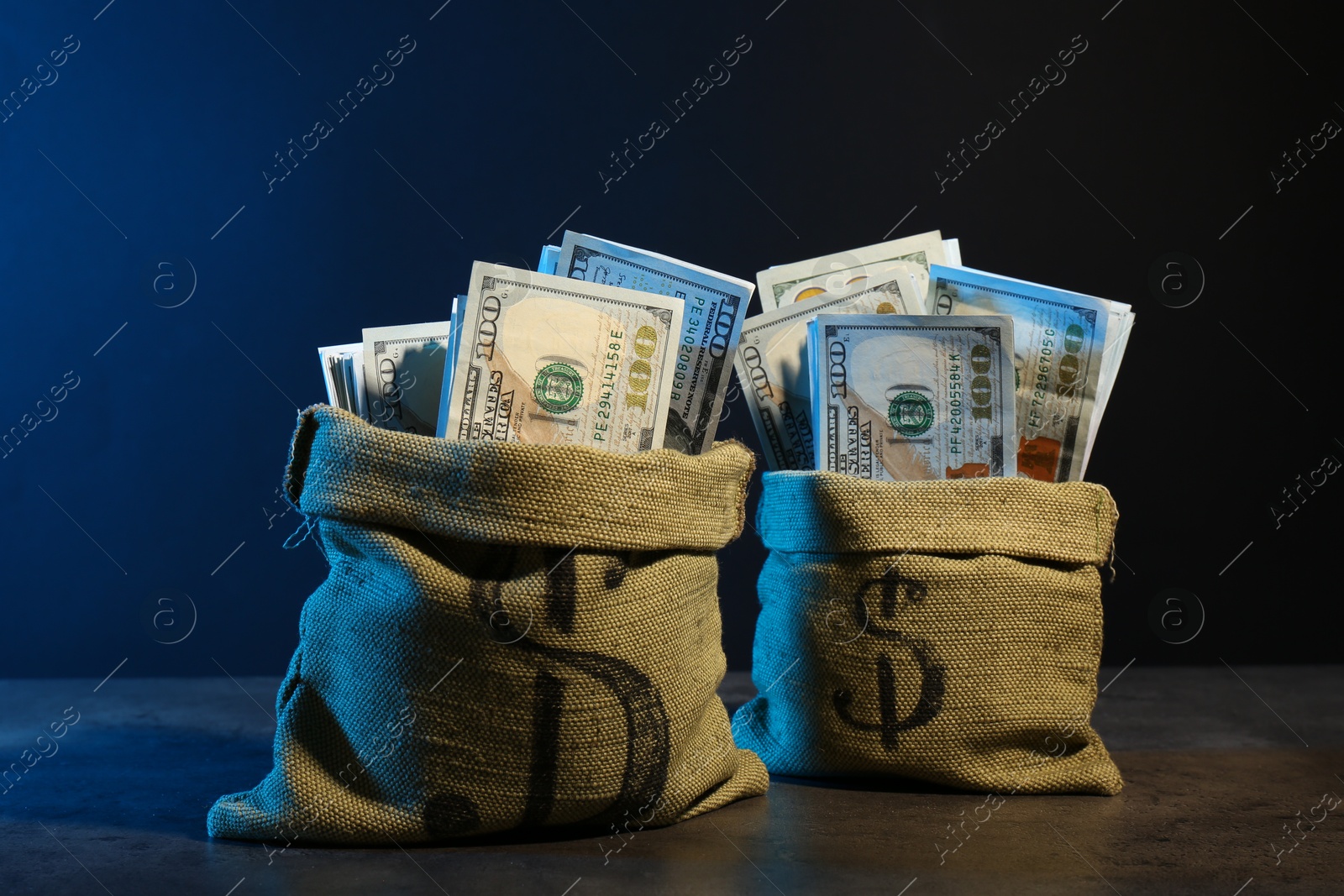 Photo of Dollar banknotes in burlap sacks on grey table against dark background in neon lights