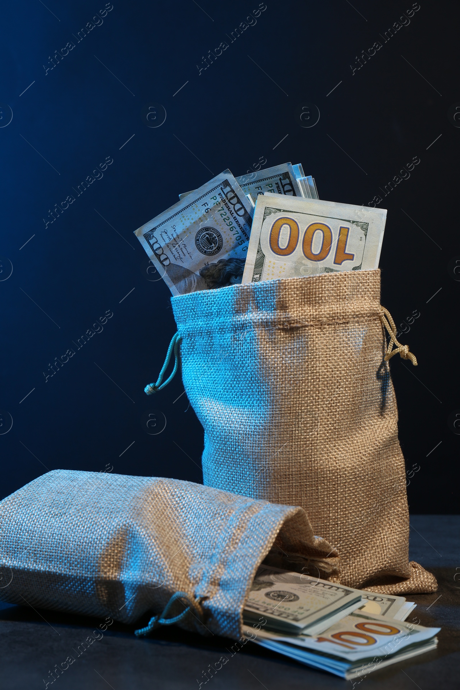 Photo of Dollar banknotes in burlap sacks on grey table against dark background in neon lights