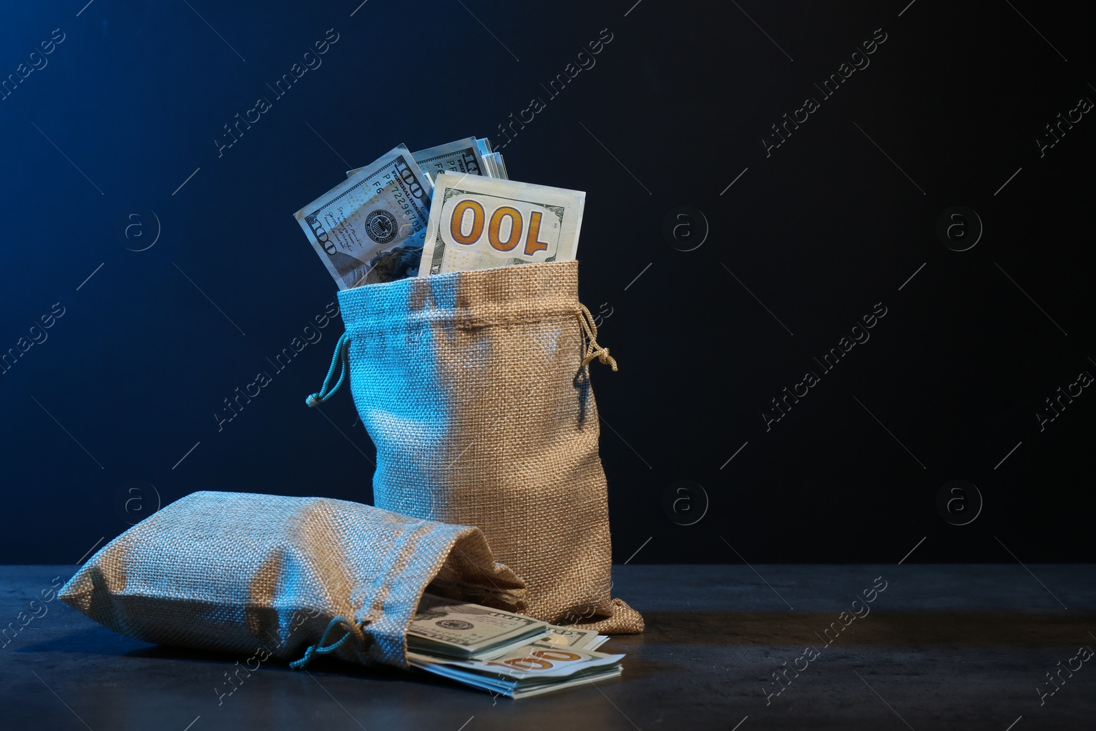 Photo of Dollar banknotes in burlap sacks on grey table against dark background in neon lights