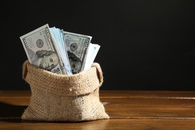 Photo of Dollar banknotes in burlap sack on wooden table against black background, space for text