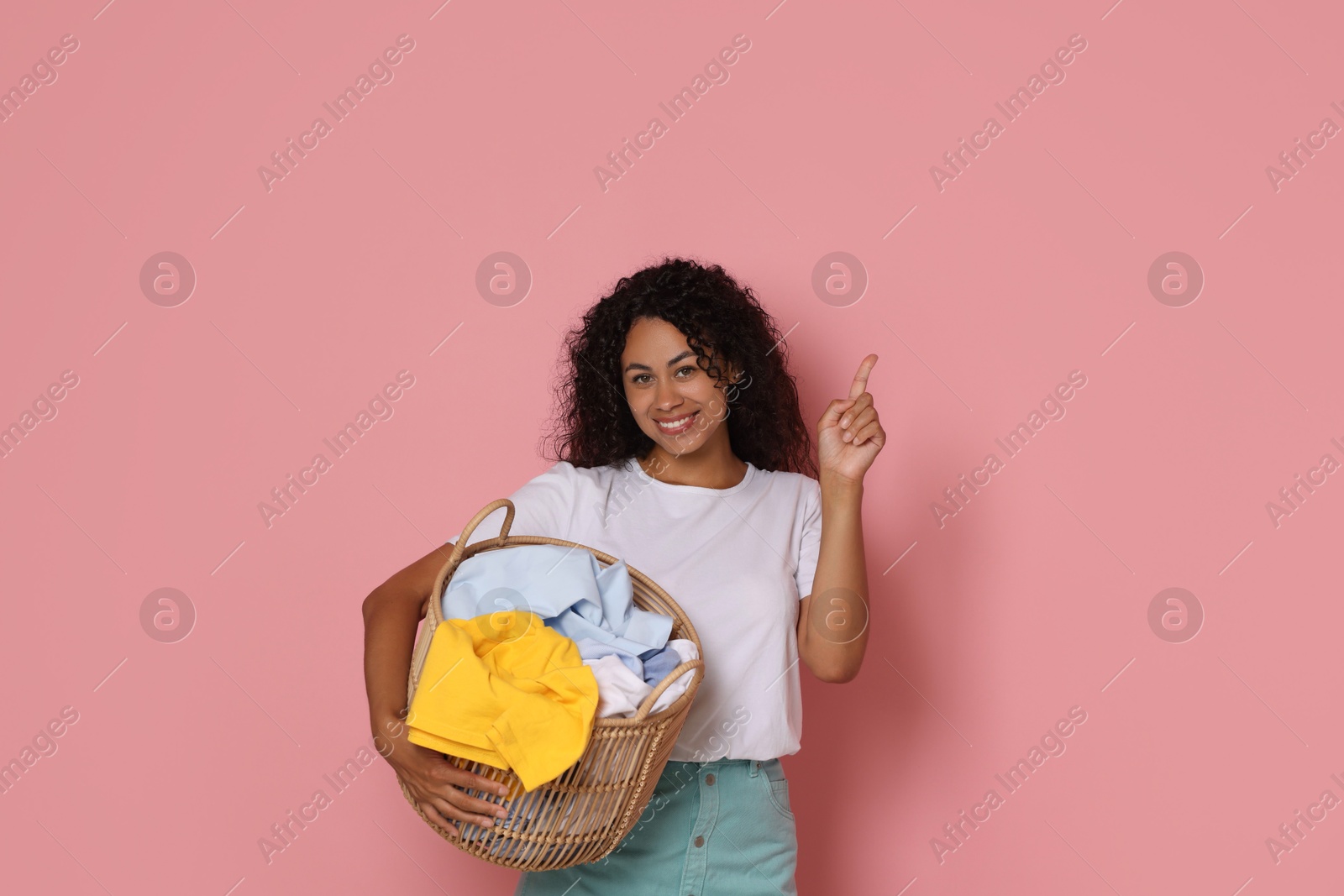 Photo of Happy woman with basket full of laundry pointing at something on pink background