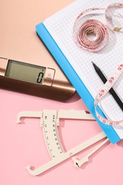 Body fat caliper, measuring tape, kitchen scale and notebook on pink background, flat lay