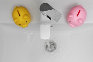 Photo of Water saving concept. Piggy banks and stream of water flowing from tap in bathroom, top view