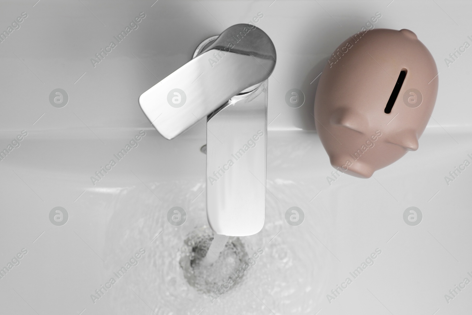 Photo of Water saving concept. Piggy bank and stream of water flowing from tap in bathroom, top view