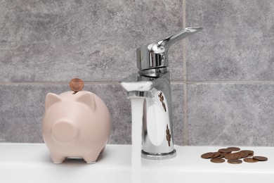 Photo of Water saving concept. Piggy bank, stream of water flowing from tap and coins in bathroom