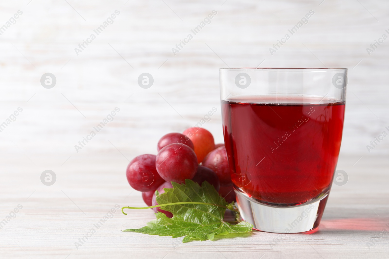 Photo of Tasty grape juice in glass, leaf and berries on light wooden table, closeup. Space for text