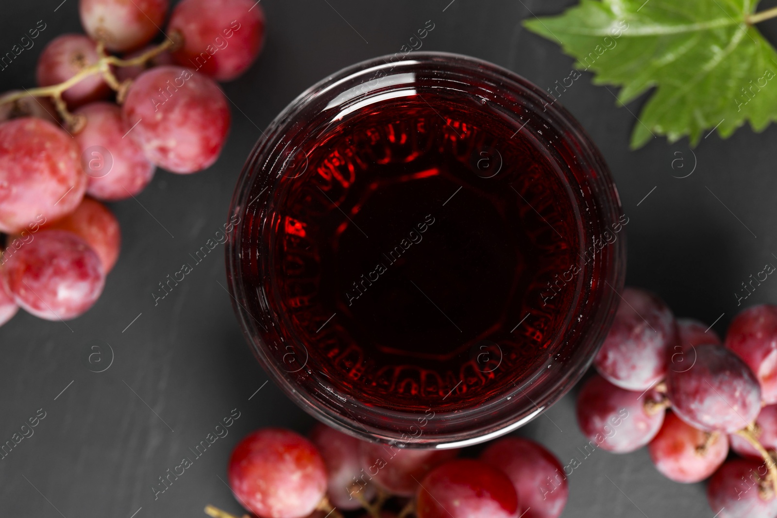 Photo of Tasty grape juice in glass, leaf and berries on dark textured table, flat lay