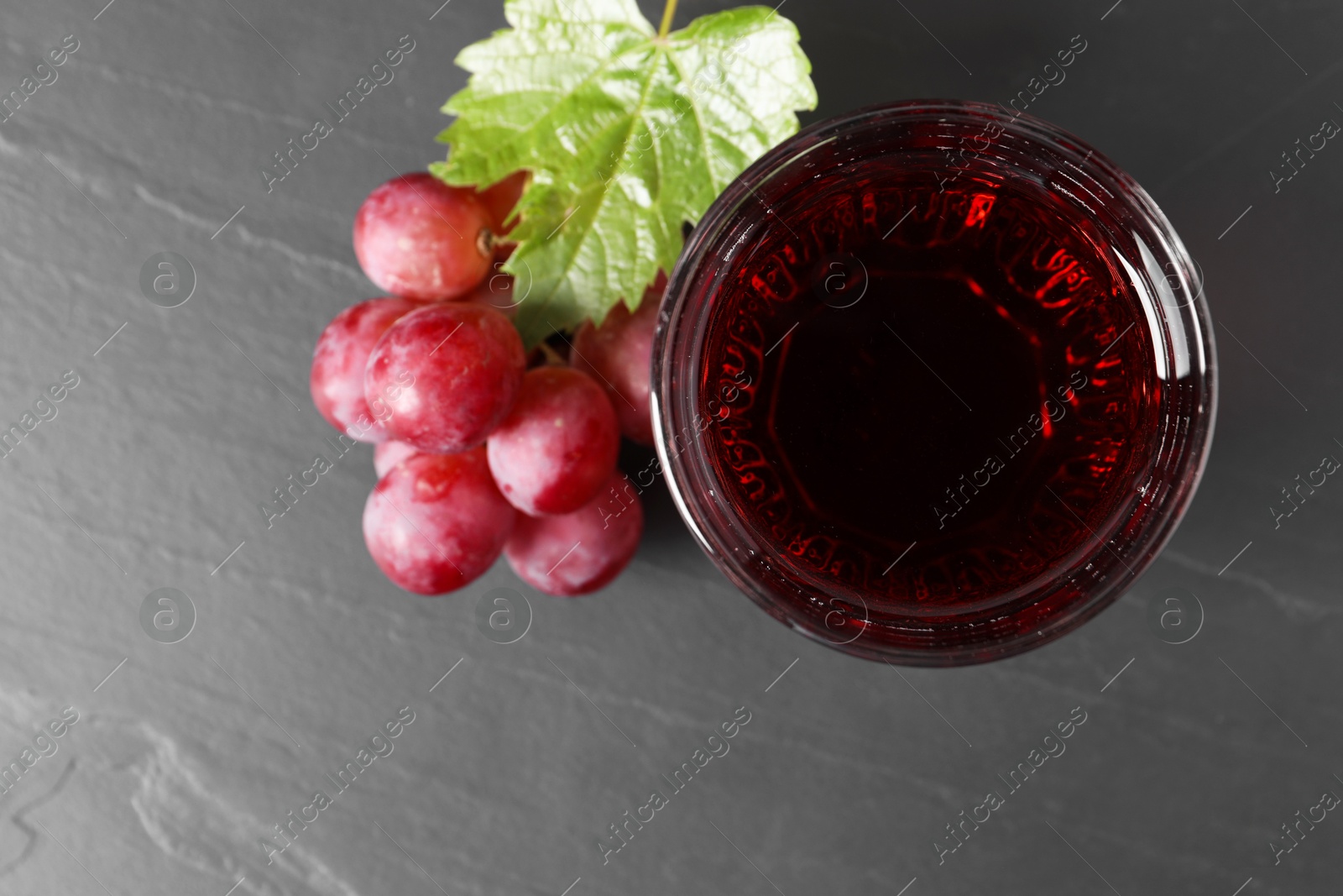 Photo of Tasty grape juice in glass, leaf and berries on dark textured table, flat lay