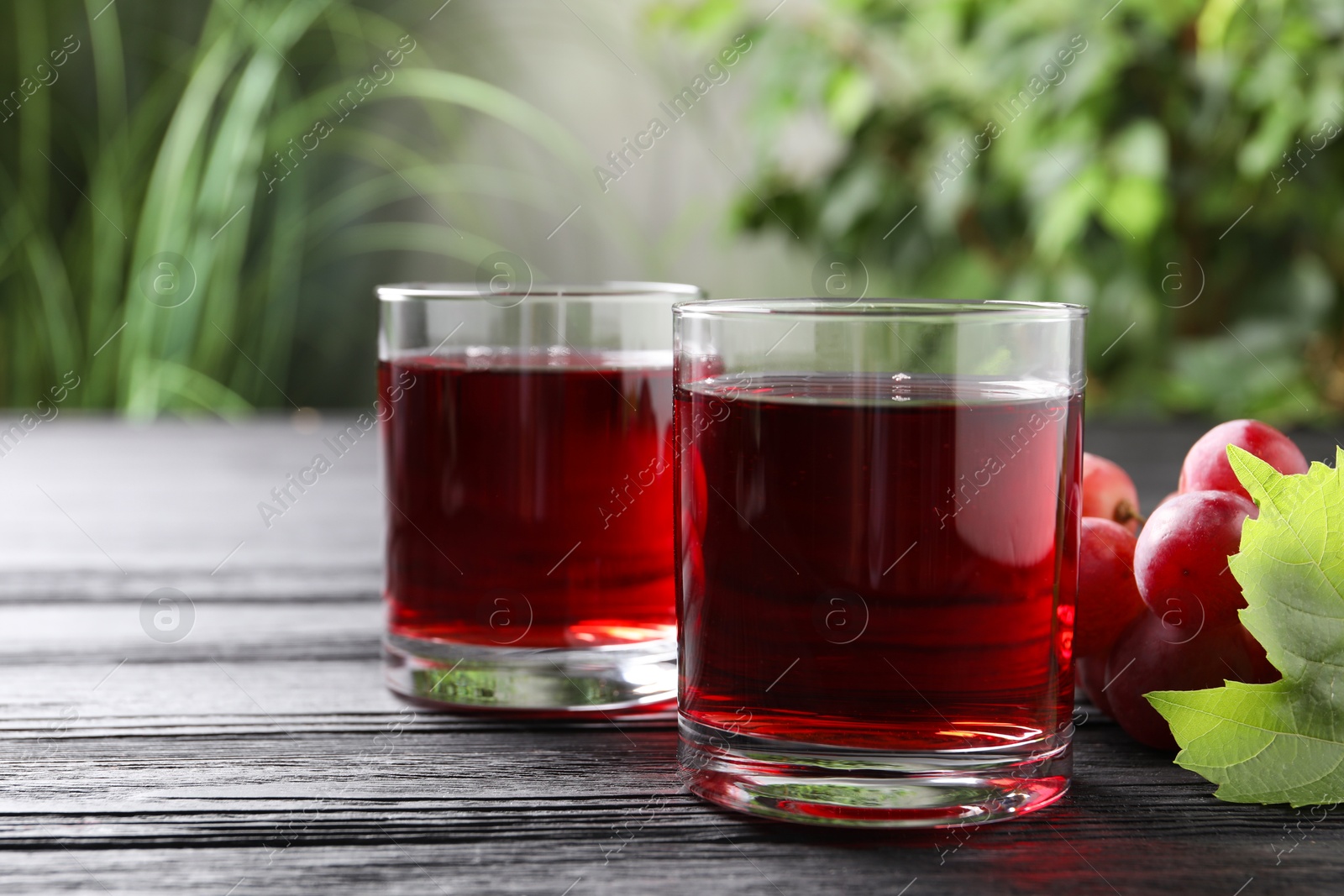 Photo of Tasty grape juice in glasses and berries on black wooden table against blurred green background, closeup