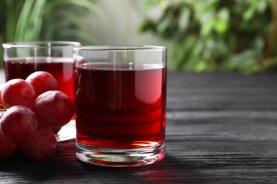 Photo of Tasty grape juice in glasses and berries on black wooden table against blurred green background, closeup. Space for text