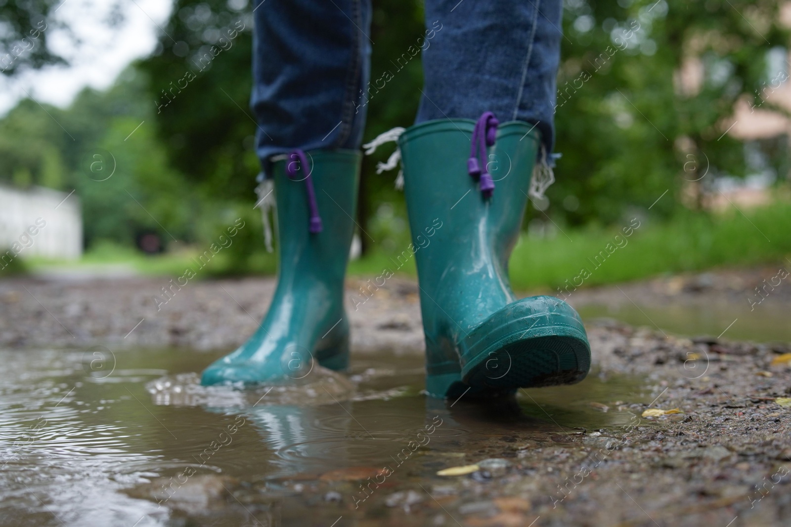 Photo of Woman wearing turquoise rubber boots walking in puddle outdoors, closeup