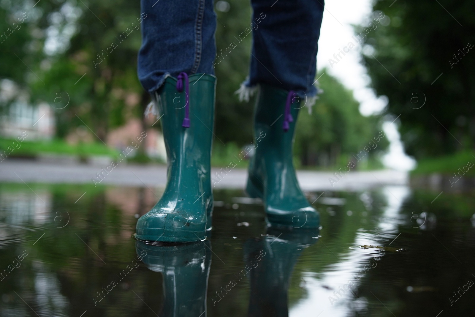 Photo of Woman wearing turquoise rubber boots walking in puddle outdoors, closeup