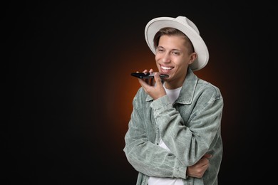 Young man recording voice message via smartphone on dark background, space for text