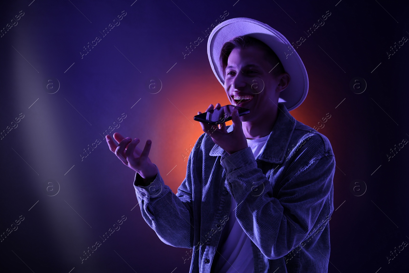 Photo of Young man recording voice message via smartphone on color background with neon lights, space for text