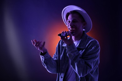 Photo of Young man recording voice message via smartphone on color background with neon lights, space for text