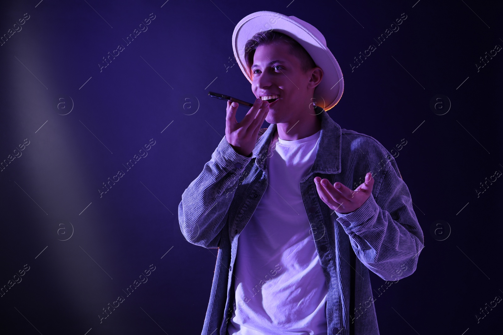 Photo of Young man recording voice message via smartphone on color background with neon lights, space for text