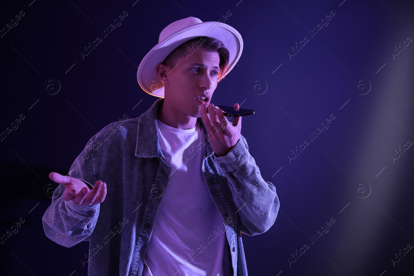 Photo of Young man recording voice message via smartphone on color background with neon lights, space for text