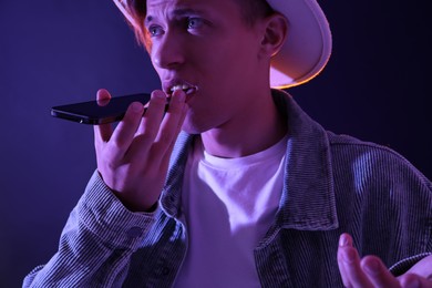 Young man recording voice message via smartphone on color background with neon lights