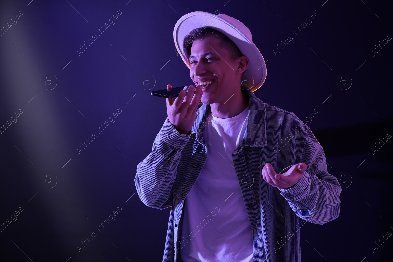 Photo of Young man recording voice message via smartphone on color background with neon lights, space for text