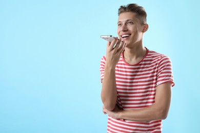 Young man recording voice message via smartphone on light blue background, space for text