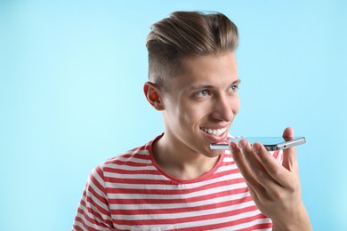 Young man recording voice message via smartphone on light blue background