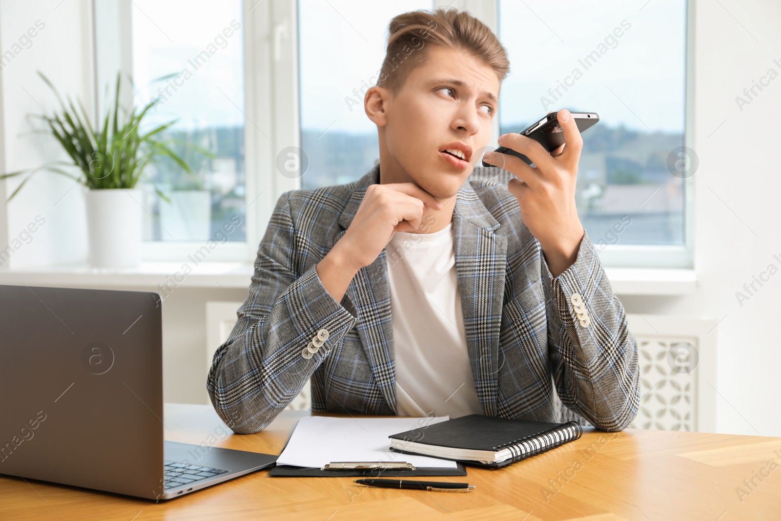 Photo of Young man recording voice message via smartphone in office