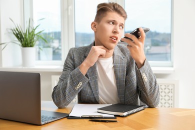 Photo of Young man recording voice message via smartphone in office