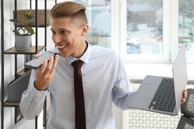 Young man with laptop recording voice message via smartphone in office