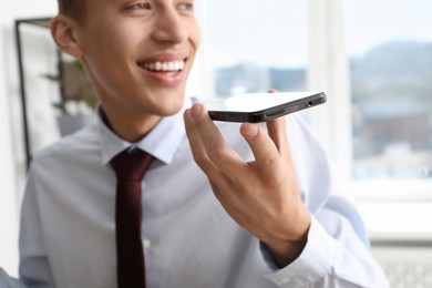 Young man recording voice message via smartphone in office, selective focus