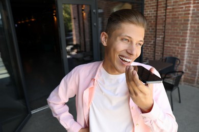 Photo of Young man recording voice message via smartphone outdoors