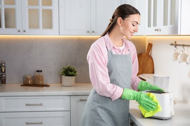 Beautiful young woman wiping toaster with rag at countertop in kitchen