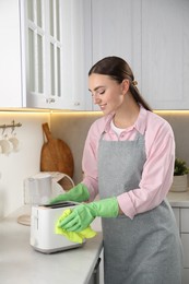 Beautiful young woman wiping toaster with rag at countertop in kitchen