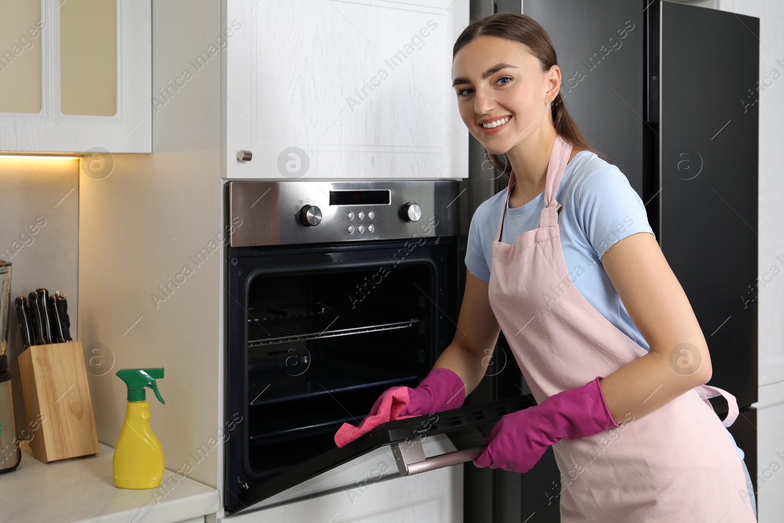 Photo of Beautiful young woman wiping electric oven with rag in kitchen