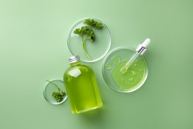 Photo of Petri dishes with cosmetic product, bottle, pipette and parsley on green background, flat lay