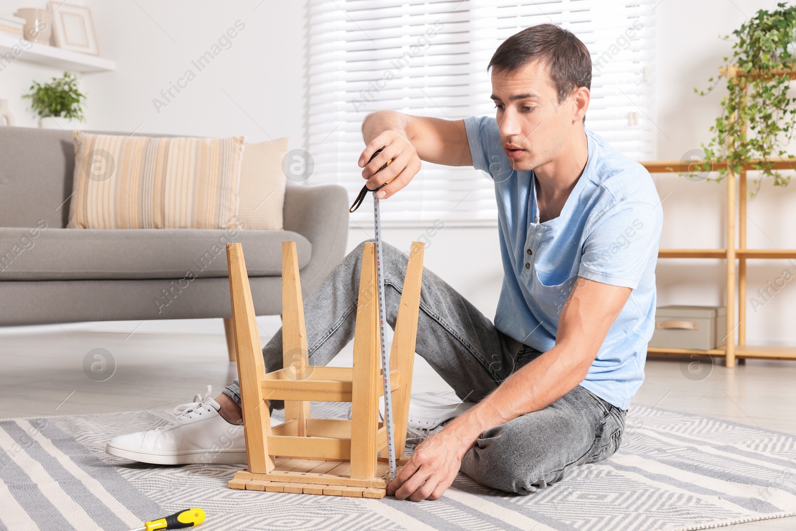 Photo of Man using tape measure while repairing wooden stool indoors