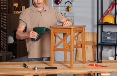 Photo of Man repairing wooden stool with electric screwdriver indoors, closeup