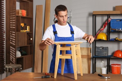 Photo of Man using tape measure while repairing wooden stool indoors