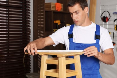 Photo of Man using tape measure while repairing wooden stool indoors