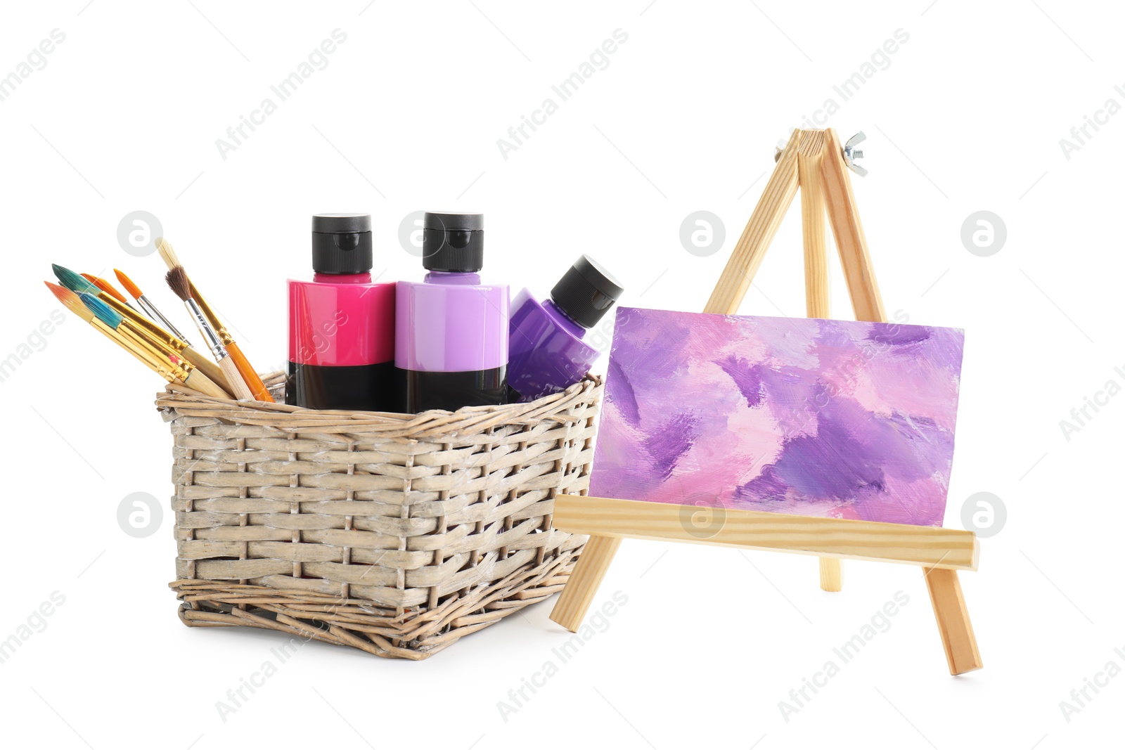 Photo of Acrylic paints of different colors with brushes in wicker basket and abstract picture on small easel against white background