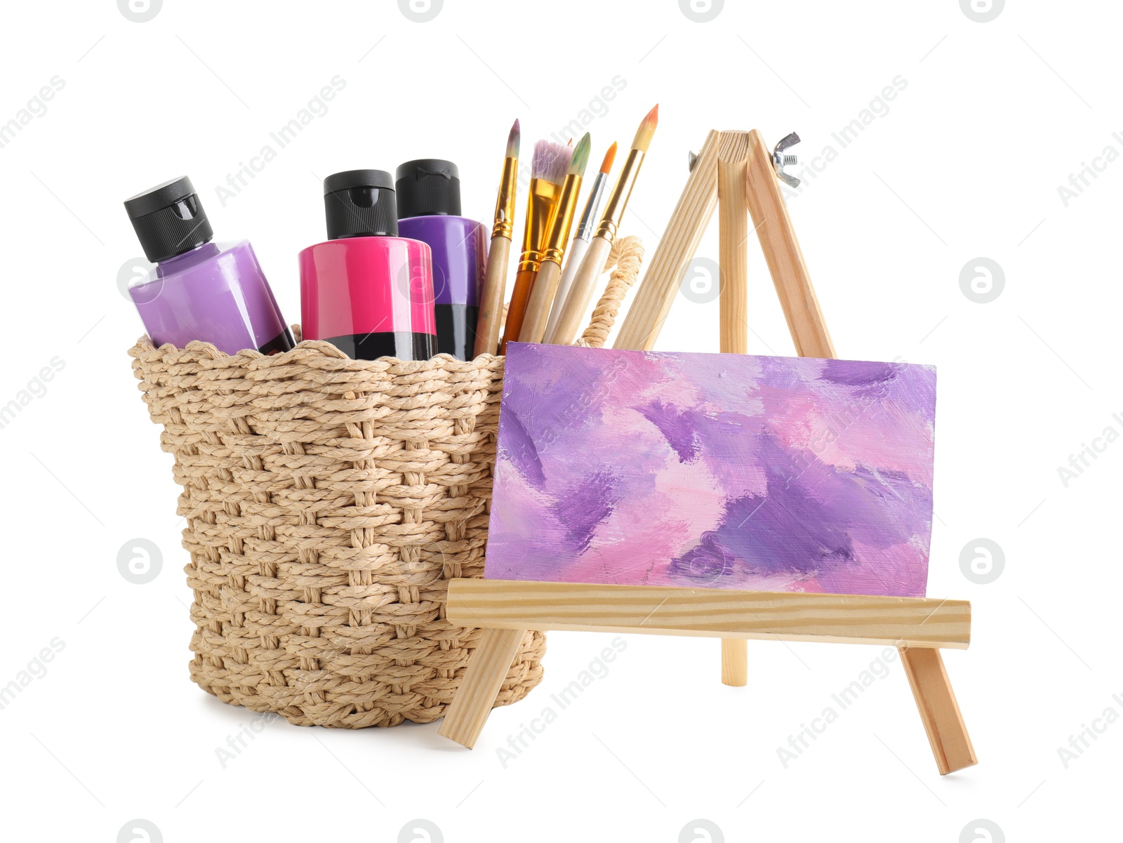 Photo of Acrylic paints of different colors with brushes in wicker basket and abstract picture on small easel against white background