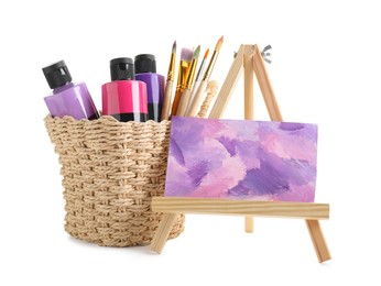 Photo of Acrylic paints of different colors with brushes in wicker basket and abstract picture on small easel against white background