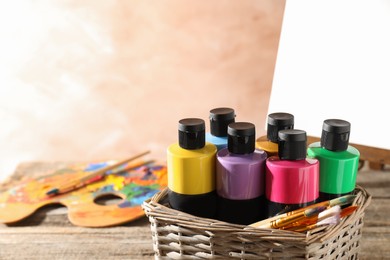 Photo of Acrylic paints of different colors and brushes in wicker basket on wooden table, space for text