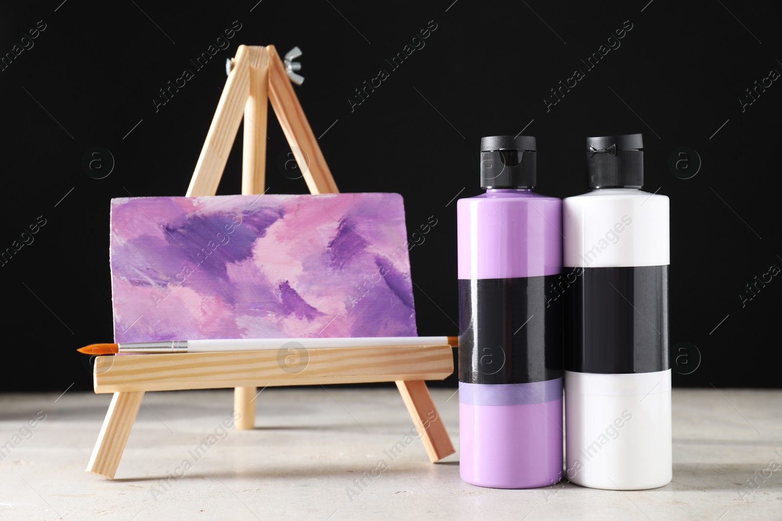 Photo of Acrylic paints of different colors in bottles, brush and small easel with abstract picture on light table