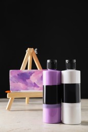 Acrylic paints of different colors in bottles, brush and small easel with abstract picture on light table